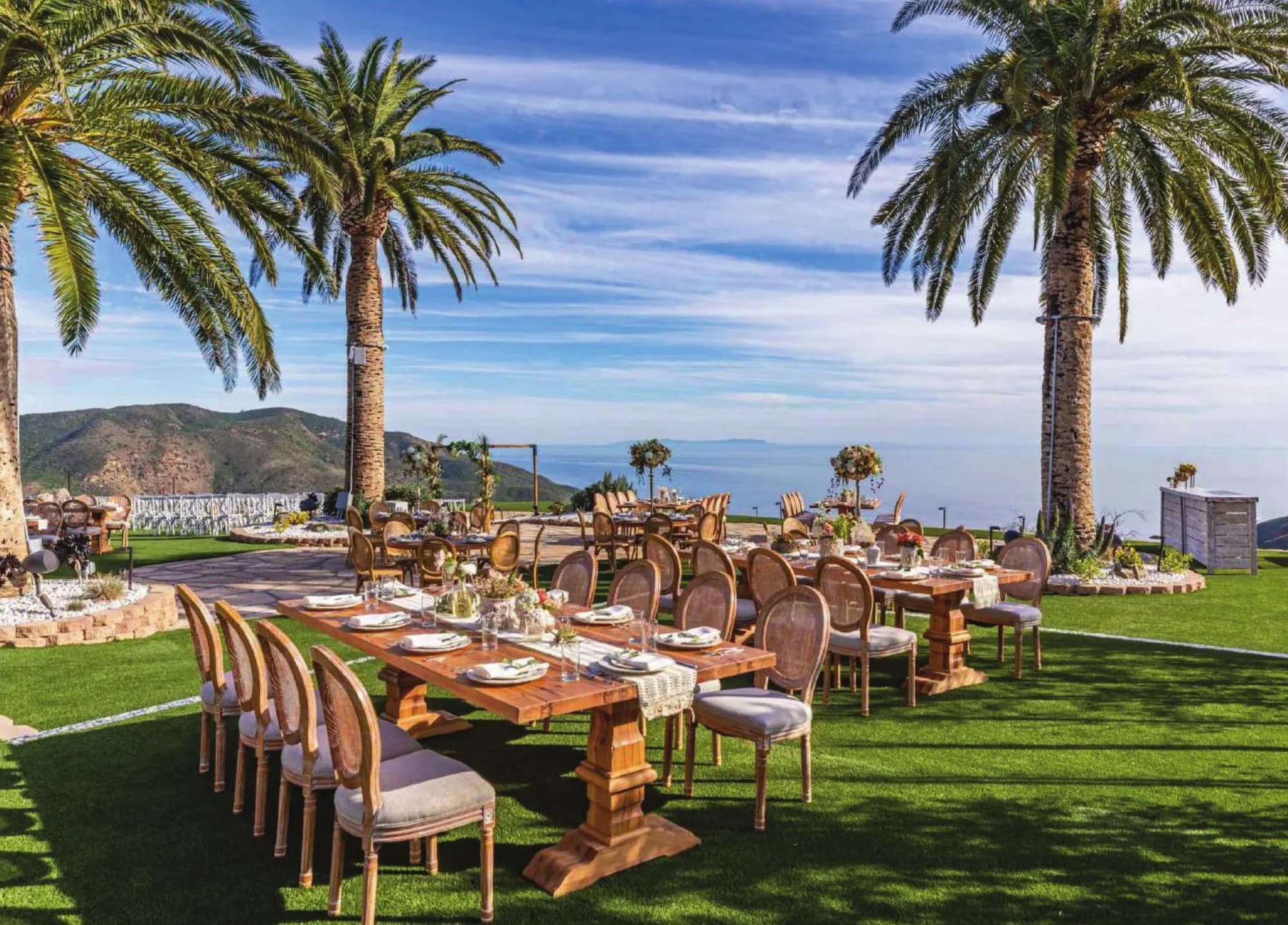 Long wooden dining tables set under palm trees from the Luxury Event Planning rental catalog